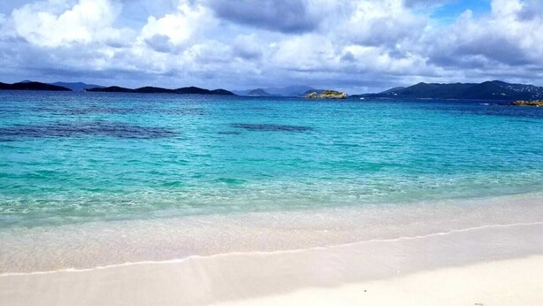 A beach with clear turquoise water, white sand, and distant mountains under a cloudy sky. - Our Walk in Paradise Level at Sapphire Village, East End (St Thomas)