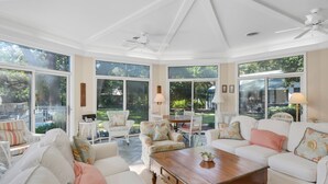 Un salón amplio con muebles blancos, ventanas grandes y un ventilador de techo.