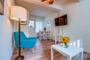 A living room with a blue chair, a white table with a vase of sunflowers, a floor lamp, a TV mounted on the wall, and a dining area in the background.