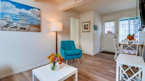 A living room with a ceiling fan, a painting of beach chairs, a teal armchair, a white table with a vase of flowers, and a dining area with a table and chairs.