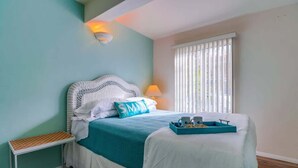A bedroom with a bed, a bedside table, a window with blinds, and a wall-mounted light fixture.