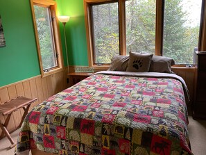 A bedroom with a large bed, a wooden nightstand, a green wall, and a window with a view of trees.