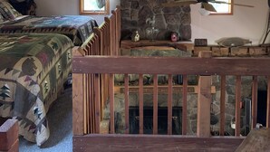 A cozy attic bedroom with a stone wall, wooden beams, a bed with a camouflage quilt, a wooden bench, and a stone fireplace. - A Rustic Retreat at Wolf Hollow Farm (Andes)