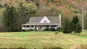A two-story house with a porch, surrounded by a grassy field and trees with autumn foliage. - A Rustic Retreat at Wolf Hollow Farm (Andes)