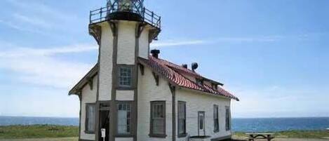 A lighthouse with a tower and a base, situated on a grassy area with a bench and a picnic table nearby.