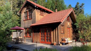 Une cabane en bois avec un toit recouvert de tuiles rouges, une terrasse en bois et un espace patio aménagé avec du mobilier d’extérieur.