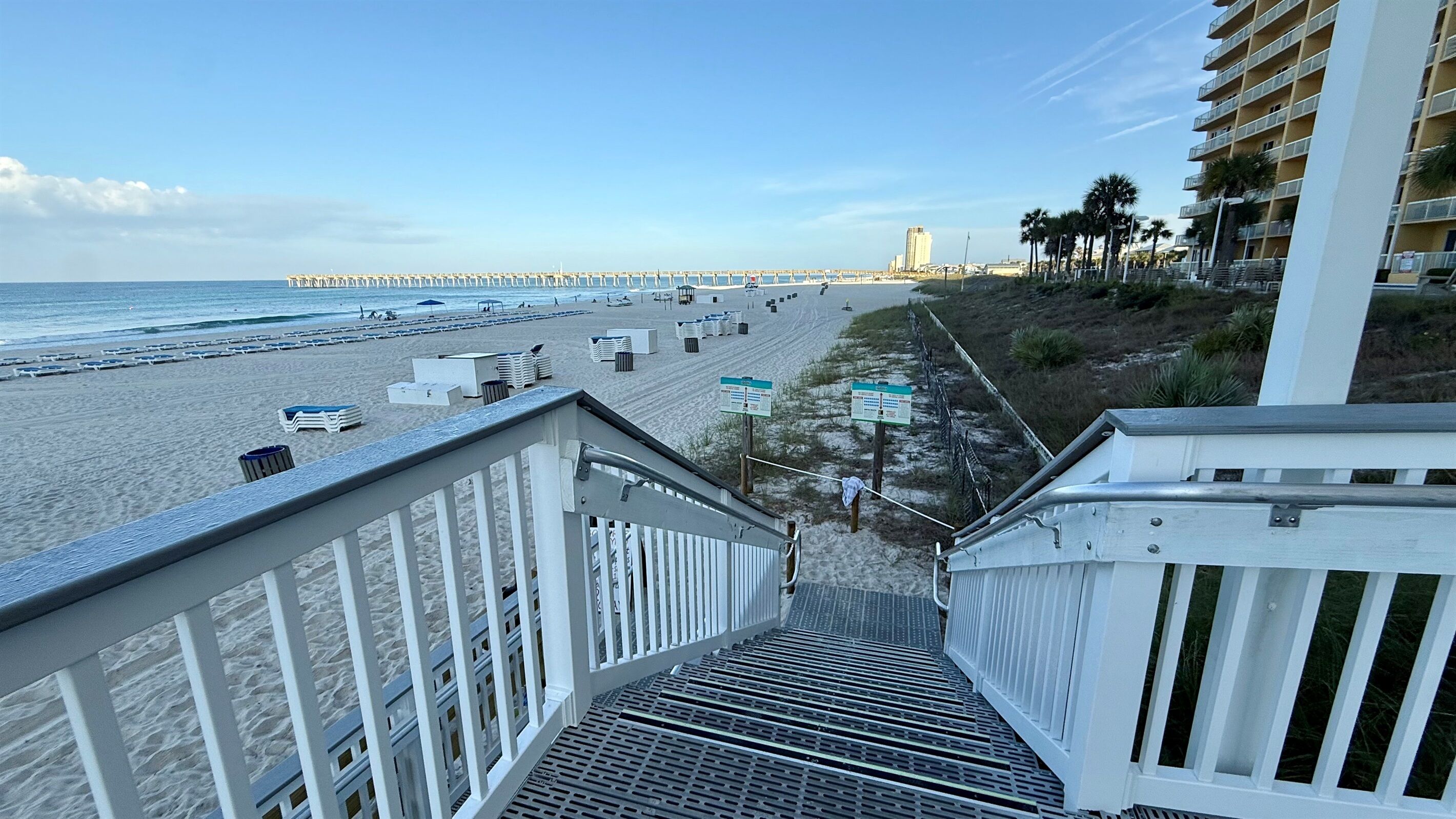 Um hotel à beira-mar com uma escadaria que desce à praia, um cais ao longe e um edifício alto.