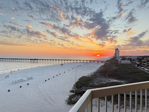 A beach with a pier extending into the water, a sunset in the background, and a building with a balcony overlooking the scene.