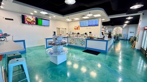 A brightly lit indoor space with a teal floor, a counter with various food items, and multiple screens displaying menus.
