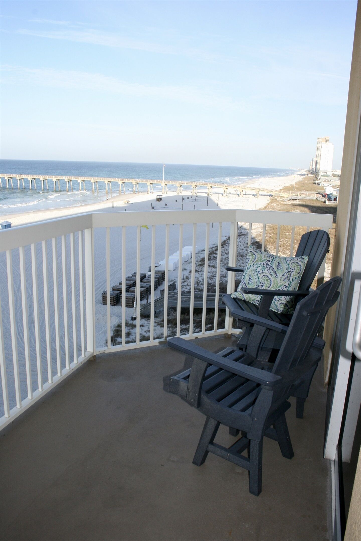 A balcony with two Adirondack chairs, one with a patterned cushion, overlooking a beach with a pier and buildings in the distance.