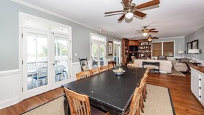A spacious dining area with a large black table surrounded by wooden chairs, a ceiling fan, and a bookshelf filled with books. - Oceanfront Home, Stunning Views, Sun Deck, Screened Porch, Walk to restaurants! (Sullivan's Island)