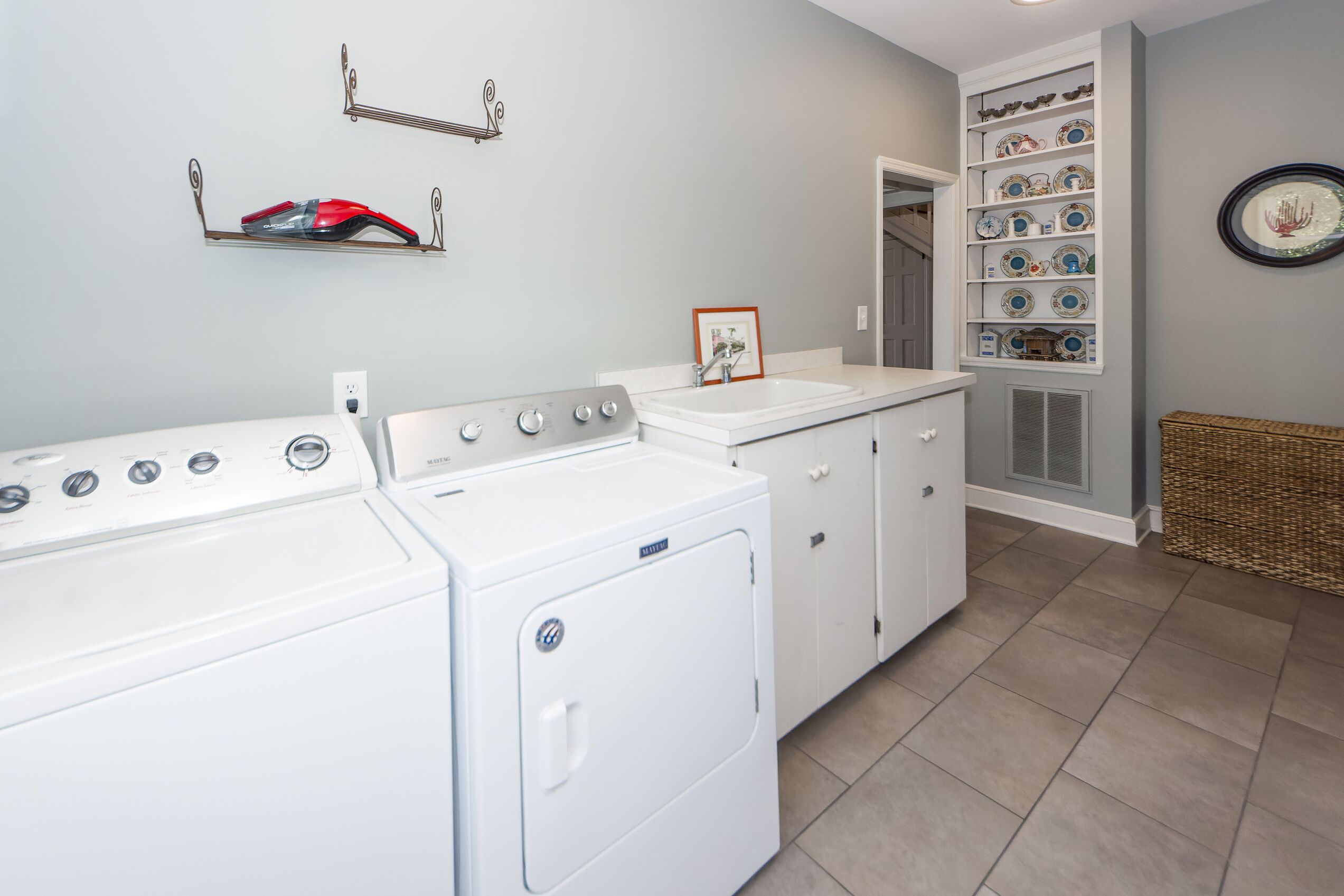 A laundry room with a washer and dryer, a sink, and a shelving unit with decorative plates.
