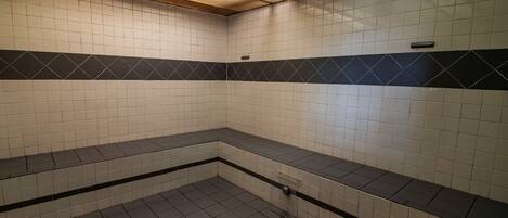 Un baño con azulejos, un banco en esquina, suelo de azulejos y techo con viga de madera.