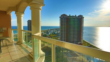 A balcony with a glass railing overlooking a coastal cityscape, featuring high-rise buildings and a view of the sea.