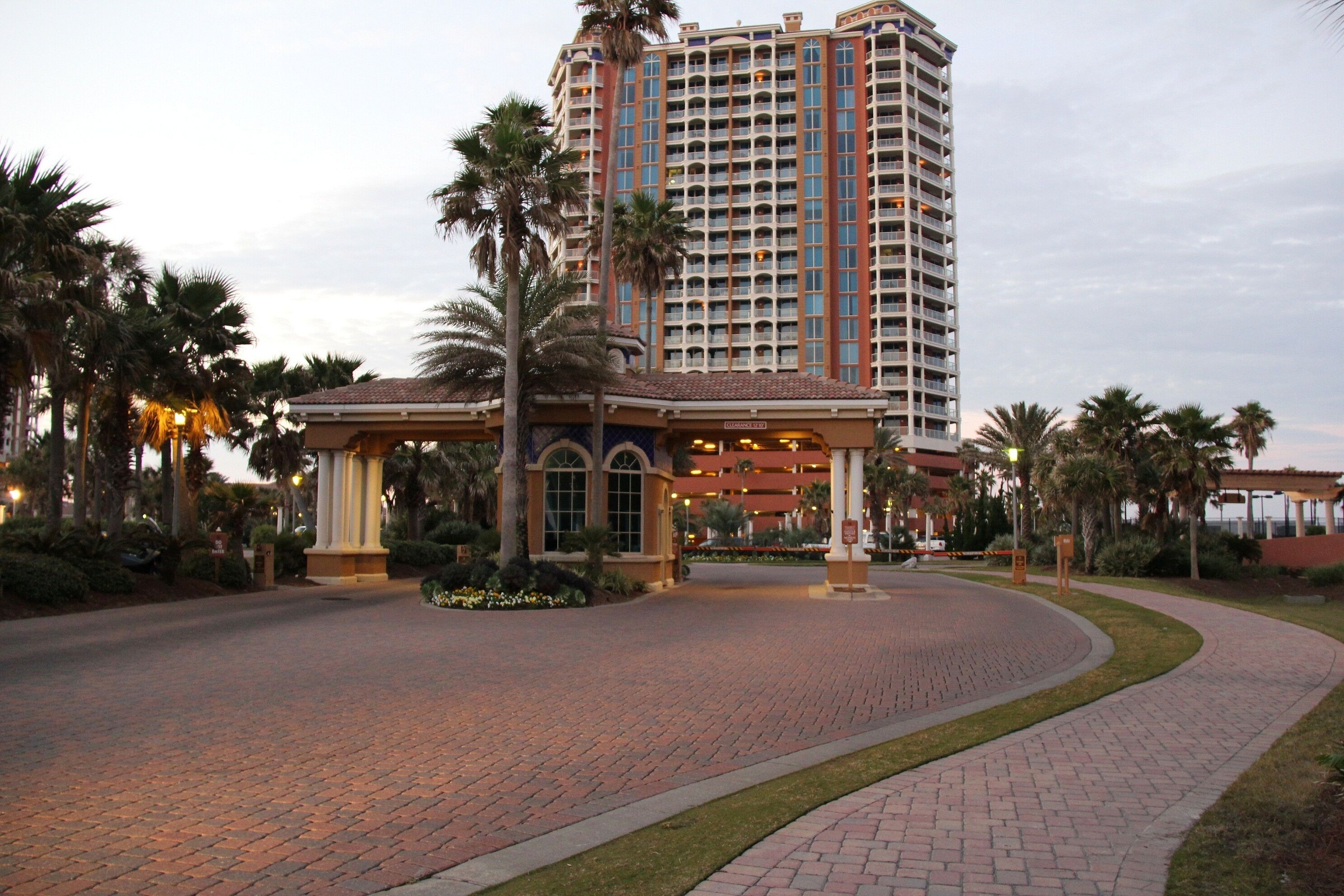 A hotel entrance with a curved brick pathway, palm trees, and a large building with multiple floors and balconies.