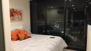 A modern bedroom with a large bed, white bedding, and orange pillows. It features a cityscape view through floor-to-ceiling glass windows.