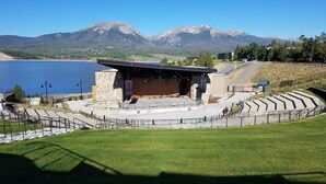 A theater with a stage and seating area, surrounded by a grassy area and a lake, with mountains in the background. - Dillon Lake 2 BR Condo-Pool, Hot Tub, 4 lift passes at Keystone-Breck & Arapahoe (Dillon)