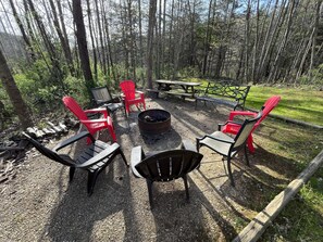 An outdoor seating area with red and black chairs, a fire pit, and a picnic table surrounded by trees.