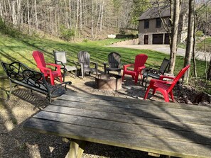 Una terraza de madera con sillas Adirondack y una fogata, rodeada de un área boscosa y una casa al fondo.