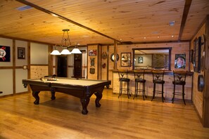 A pool table with four chairs, a bar area with stools, wooden walls, and a wooden ceiling.