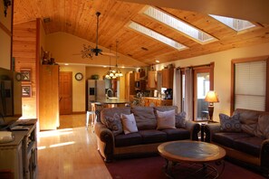 A spacious living room with a high ceiling and skylights, a cozy seating area with a sofa and armchairs, and a kitchen in the background. - Harvest Moon Lodge at Harvest Moon Cottages, LLC (Rockbridge)