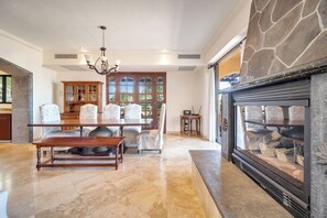 A spacious living room with a fireplace, a dining area with a wooden table and chairs, and a large window with a view of trees.