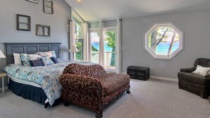 A bedroom with a bed, a patterned armchair, a window with a view, and a ceiling fan.