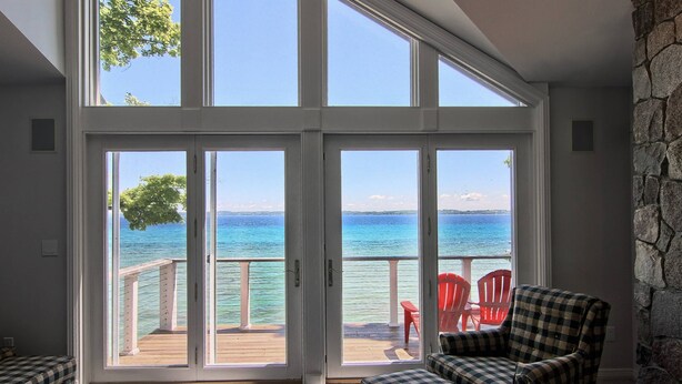 A room with a large sliding glass door leading to a balcony with a view of the ocean. There are two checkered armchairs and a red chair on the balcony.