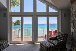 A room with a large sliding glass door leading to a balcony with a view of the ocean. There are two checkered armchairs and a red chair on the balcony. - Beautiful Large House On Water Just 5 Miles To TC (Traverse City)