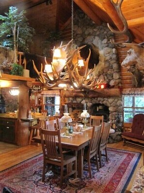 A rustic lodge interior with a stone fireplace, wooden dining table, and antler chandeliers. - Four Seasons Log Guest House (Gallatin Gateway)