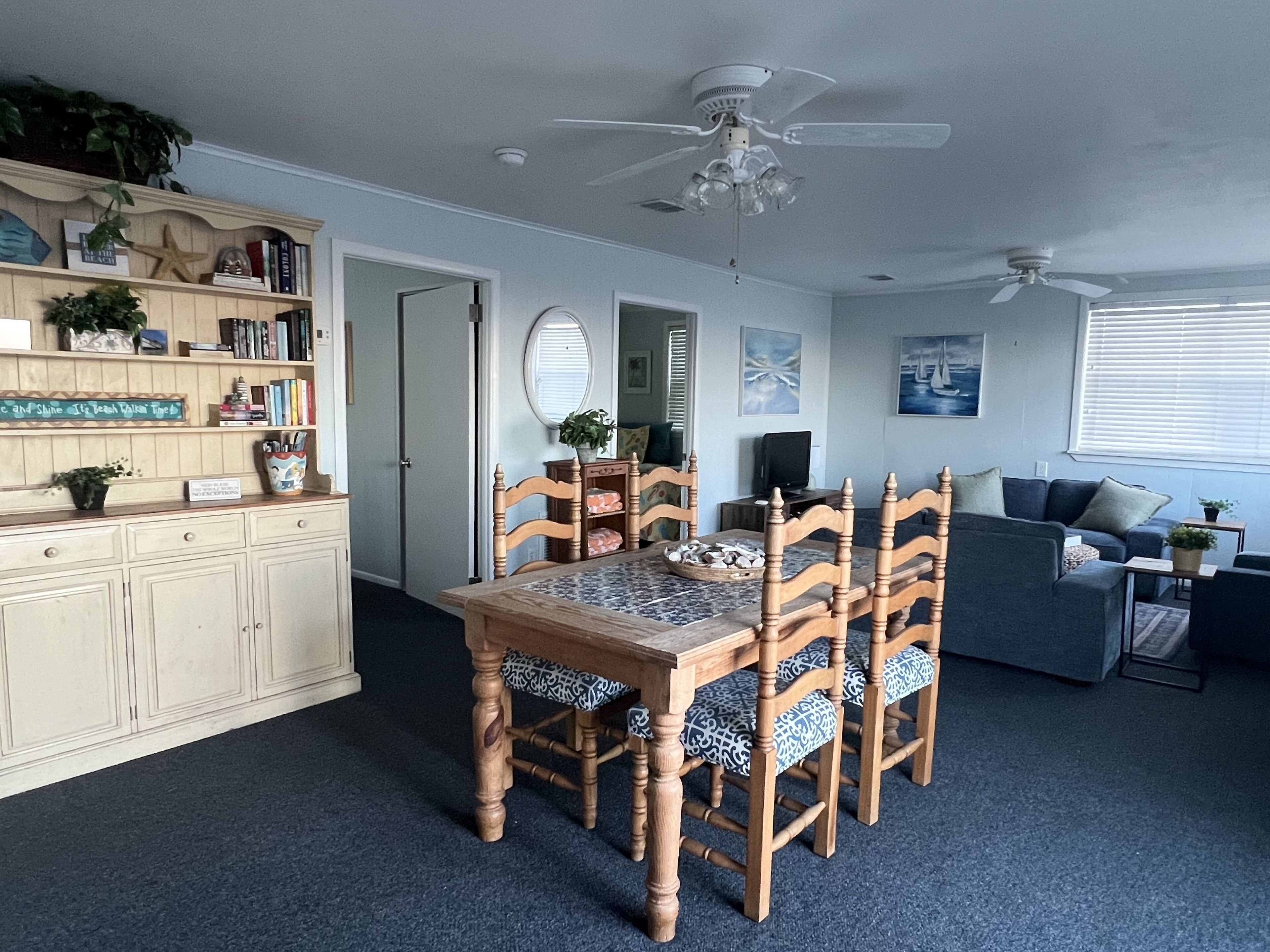 A dining area with a wooden table and chairs, a ceiling fan, a bookshelf, and a seating area with a sofa and armchairs.