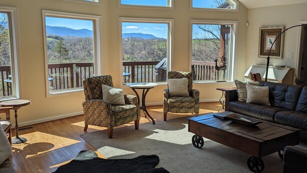 Ein geräumiges Wohnzimmer mit großen Fenstern und Blick auf die Berge, eine gemütliche Sitzecke mit gemusterten Sesseln, ein Ledersofa und ein rustikaler Couchtisch.