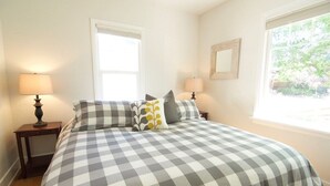 A bedroom with a checkered bedspread, two bedside tables with lamps, a window with a view of greenery, and a mirror on the wall.