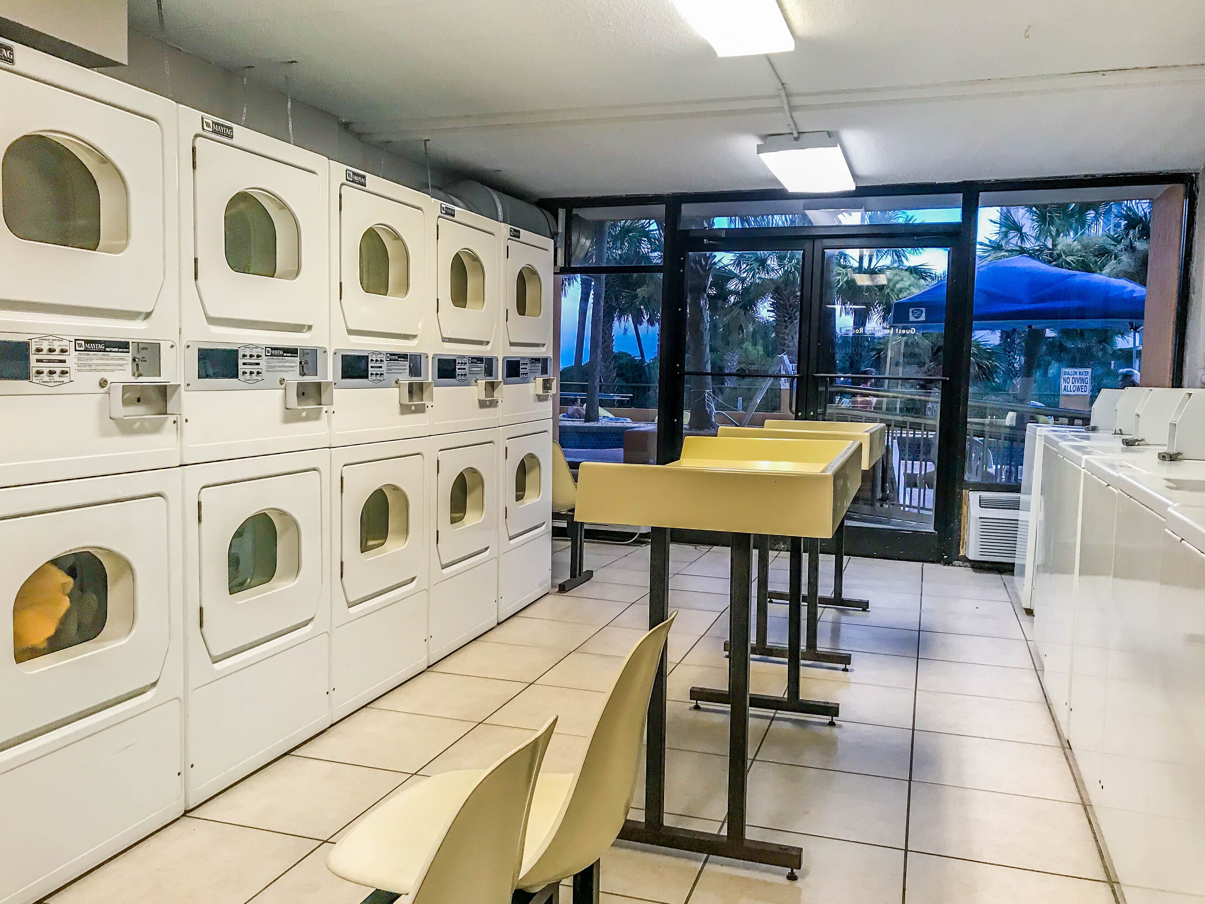 A laundromat with multiple washing machines, chairs, and a view of palm trees outside.