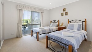 A bedroom with two single beds, a large window with curtains, a small bedside table with a lamp, and a wall-mounted picture. - Resort Style Apartment (Ground Floor) - Pool & More. Pets Welcome. (Lakes Entrance)