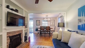 A living room with a fireplace, a TV, a sofa, and a dining area with a chandelier. - Quintessential Cottage near beaches and Newport center. (Middletown)