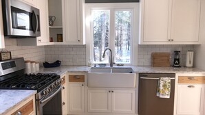 Una cocina con gabinetes blancos, estufa de acero inoxidable, microondas, pileta y ventana con vista a árboles.