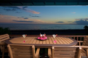 Een houten tafel met twee martini glazen en een schaal roze bloemen, voor een achtergrond van een zonsondergang boven de oceaan.