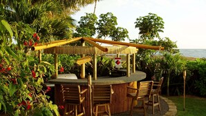 Zona de bar al aire libre con techo de paja, rodeada de sillas y una piscina.