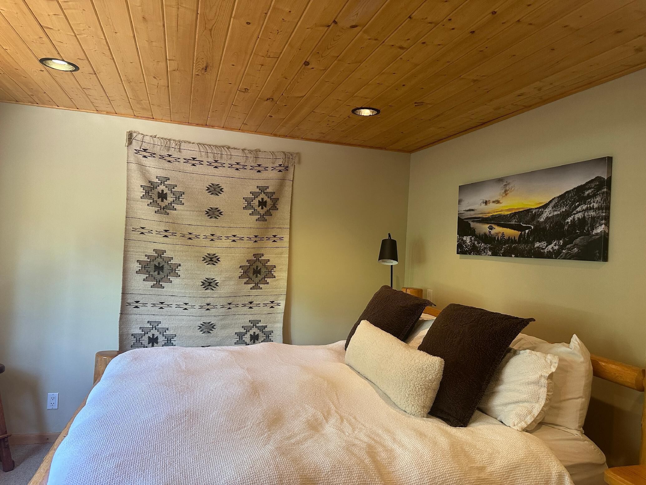 A bedroom with a wooden ceiling, a bed with multiple pillows, a wall-mounted lamp, and a landscape painting.