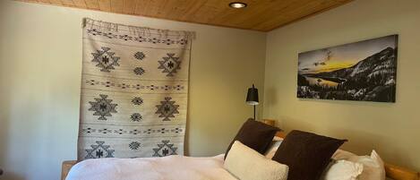A bedroom with a wooden ceiling, a bed with multiple pillows, a wall-mounted lamp, and a landscape painting.