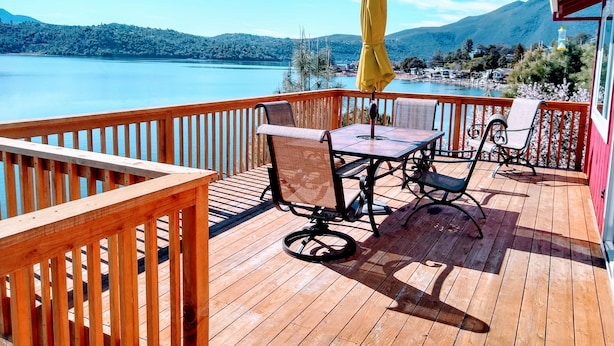 Een houten terras met een tafel en stoelen, uitkijkend op een meer en bergen.