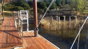 Een houten terras met een metalen hekwerk, met uitzicht op een waterpartij, omgeven door begroeiing en gebouwen.