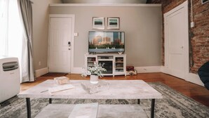Una sala de estar con televisor de pantalla plana, un mueble blanco para equipo electrónico, una mesa de piedra y una pared de ladrillo.