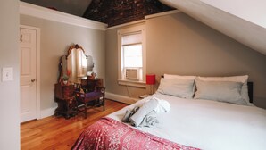 Un dormitorio con techo inclinado, cama con ropa de cama blanca, lámpara roja, cómoda, silla y ventana con persianas.