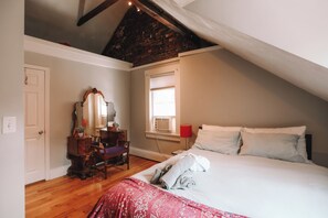 Un dormitorio con techo inclinado, cama con ropa de cama blanca, lámpara roja, cómoda, silla y ventana con persianas.