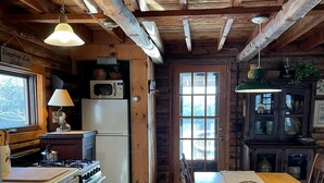 A rustic kitchen with wooden beams, a stove, a refrigerator, and a dining table with chairs. - Charming Log Cabin on Private Pond Nestled in Woods Near Holiday Valley Resort 
 (Great Valley)
