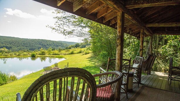 A wooden deck with wicker chairs overlooking a lake and forested hills. - Charming Log Cabin on Private Pond Nestled in Woods Near Holiday Valley Resort 
 (Great Valley)