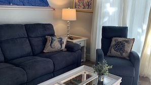 A living room with a dark blue sofa, a white coffee table, and a blue armchair. There is a painting on the wall and a lamp on a side table.