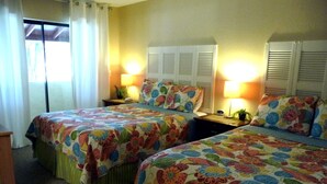A hotel room with two beds, each with floral-patterned bedding. There are two bedside tables with lamps and a small plant on one.